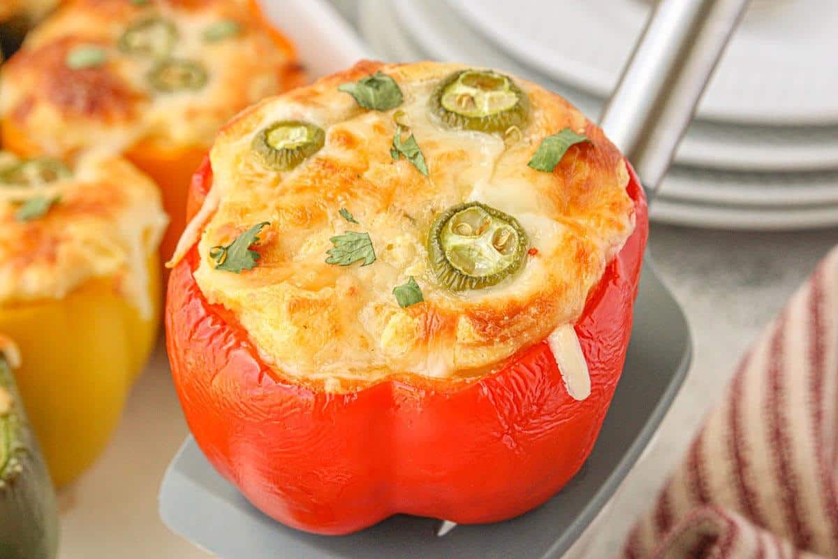 A baked red stuffed pepper topped with golden cornbread, melted cheese, and sliced jalapeños, lifted from the baking dish on a spatula and ready to serve.