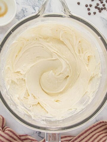 A glass mixing bowl filled with creamy white vanilla frosting on a marble countertop, surrounded by small bowls of ingredients and a red striped kitchen towel.
