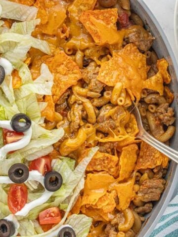 Large skillet filled with cheesy taco hamburger helper topped with lettuce, sour cream drizzle, diced tomatoes, and sliced black olives.