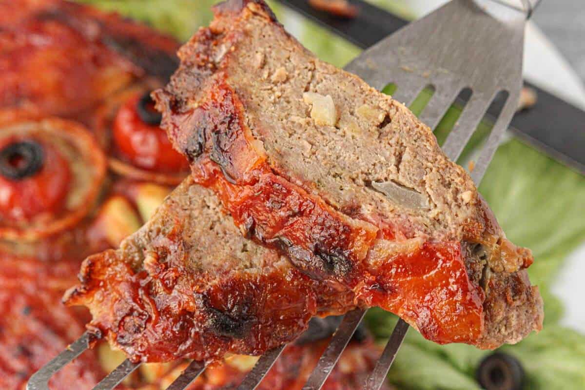 Close-up of a sliced piece of mummy meatloaf showing the moist, flavorful interior and ketchup-glazed bacon crust.