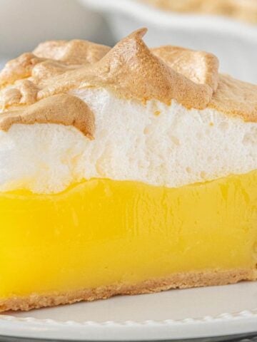 A slice of Lemon Jello Meringue Pie with a golden crust, silky lemon filling, and fluffy toasted meringue topping on a white plate with lemon curls.