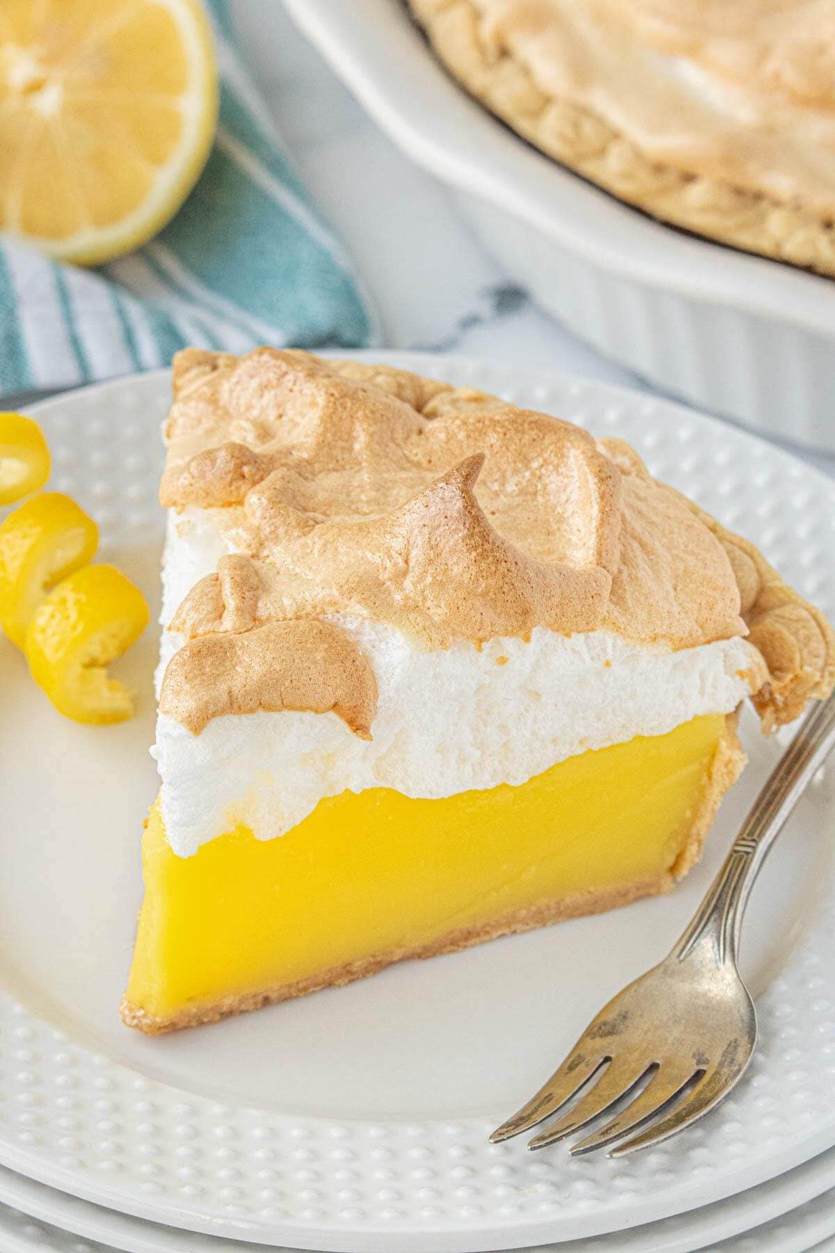 Close-up of a slice of Lemon Jello Meringue Pie showing the smooth, glossy lemon layer topped with lightly browned meringue. A lemon curl and fork is on the side of the plate.