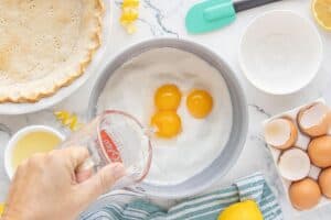 A hand pours water into a saucepan filled with sugar and egg yolks to begin the lemon filling, surrounded by lemons, eggshells, and a baked pie crust.