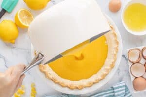 Warm lemon filling being poured into a baked pie crust from a saucepan, with lemons and eggs on the counter.