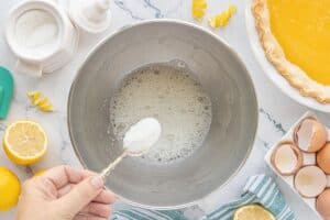 Sugar being added to whipped egg whites in a metal mixing bowl to make fluffy meringue for the pie topping.