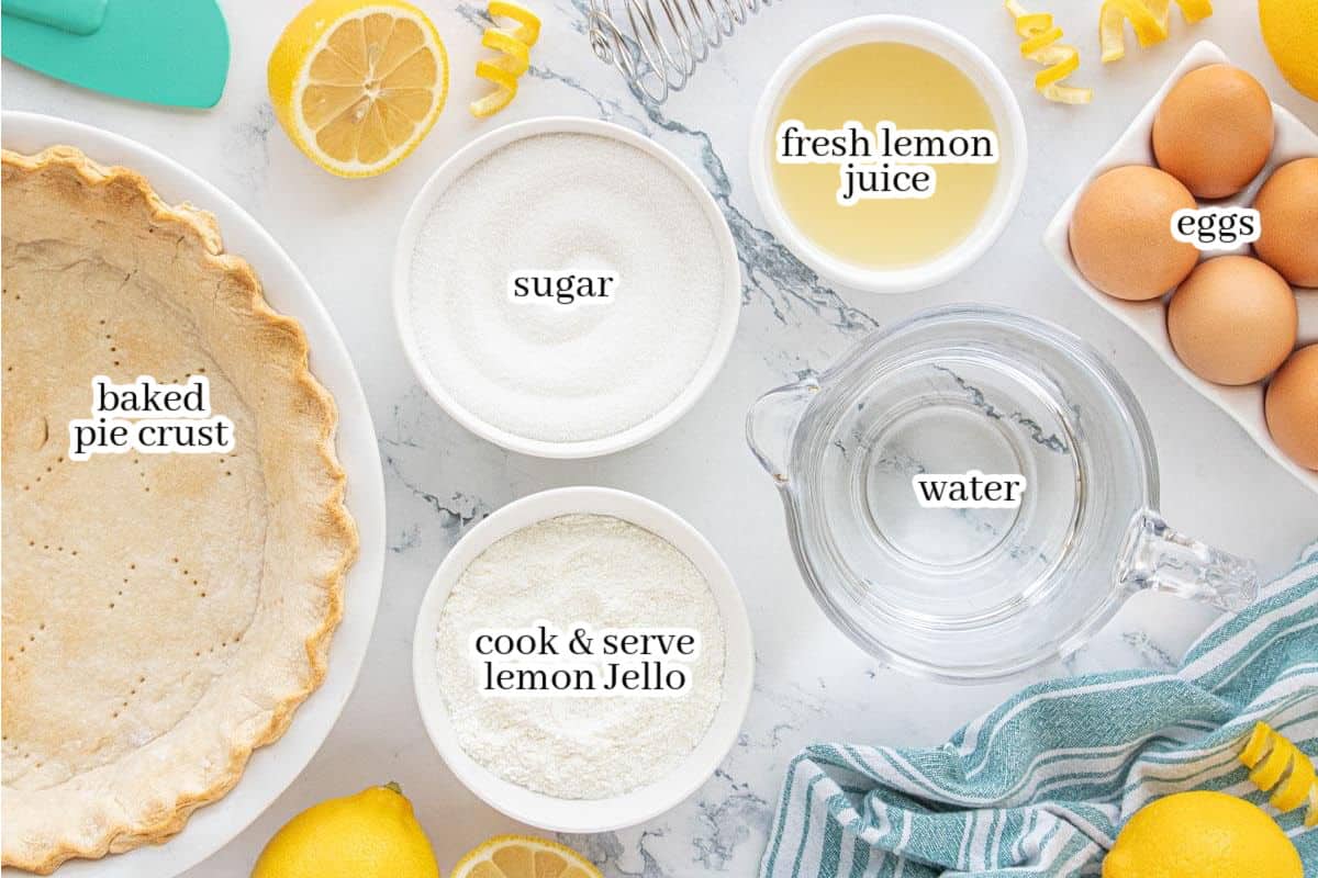 Overhead view of Lemon Jello Meringue Pie ingredients on a marble counter, including a baked pie crust, sugar, lemon juice, eggs, lemon Jello mix, and water, with lemons and a striped kitchen towel nearby.