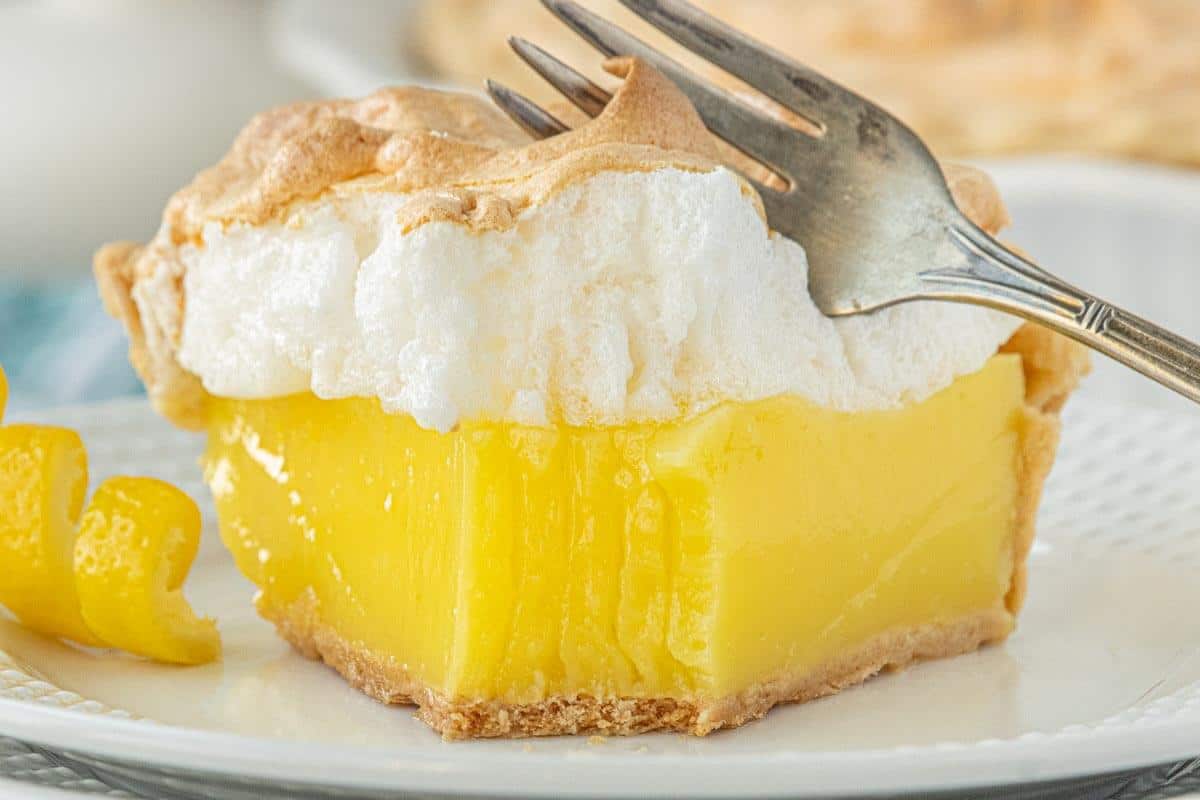 A slice of Lemon Jello Meringue Pie on a white plate with a fork, showing the bright yellow filling and toasted meringue peaks.