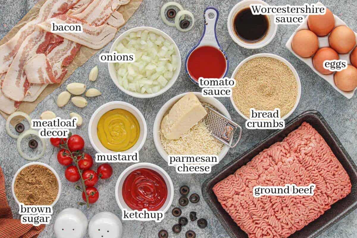 Overhead view of labeled ingredients for Mummy Meatloaf, including ground beef, bacon, eggs, breadcrumbs, onions, garlic, Parmesan cheese, ketchup, mustard, tomato sauce, Worcestershire sauce, brown sugar, and decorative items for the mummy face.