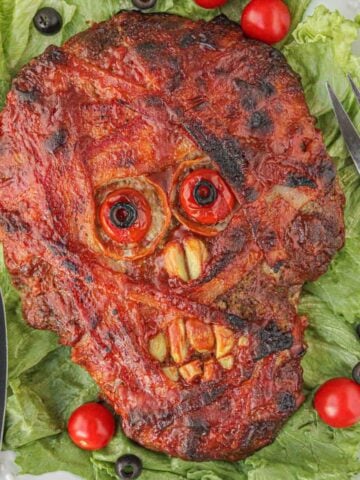 Halloween mummy meatloaf shaped like a spooky face, wrapped in bacon and baked until golden, served on platter surrounded by lettuce with cherry tomatoes and olives.