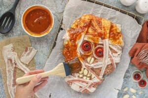 Hand brushing ketchup glaze over bacon strips on the mummy meatloaf before baking, with ingredients arranged around the tray.