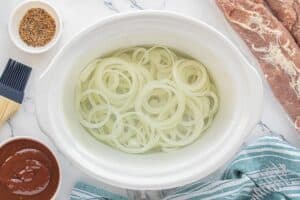 Sliced onions arranged in the bottom of a white slow cooker surrounded by barbecue sauce, seasoning blend, and pork tenderloin.