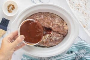 Seasoned pork tenderloins in a slow cooker with barbecue sauce being poured over the top.