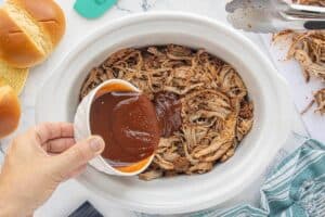 Barbecue sauce being poured over shredded pork tenderloin in a slow cooker.