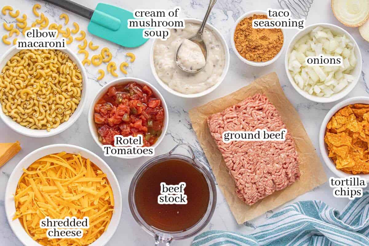 Overhead view of Crunchy Hamburger Helper ingredients including ground beef, elbow macaroni, Rotel tomatoes, cream of mushroom soup, taco seasoning, onions, beef broth, shredded cheddar cheese, and crushed tortilla chips arranged on a marble counter.
