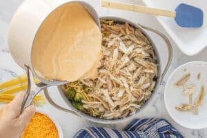 Creamy cheese sauce being poured from a pot over a mixture of cooked spaghetti, shredded chicken, and broccoli in a large pan.