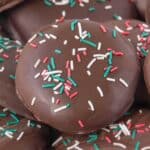 Close-up of a chocolate-covered peppermint patty topped with red, green, and white sprinkles, highlighting the shiny chocolate surface and festive decoration.