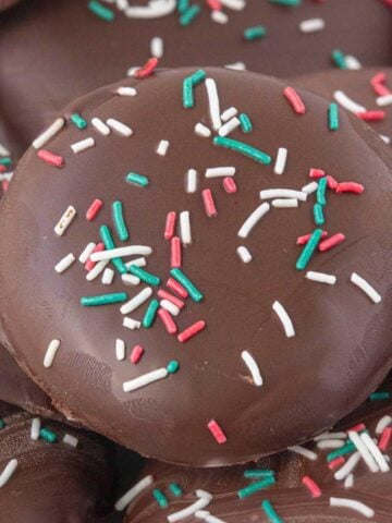 Close-up of a chocolate-covered peppermint patty topped with red, green, and white sprinkles, highlighting the shiny chocolate surface and festive decoration.