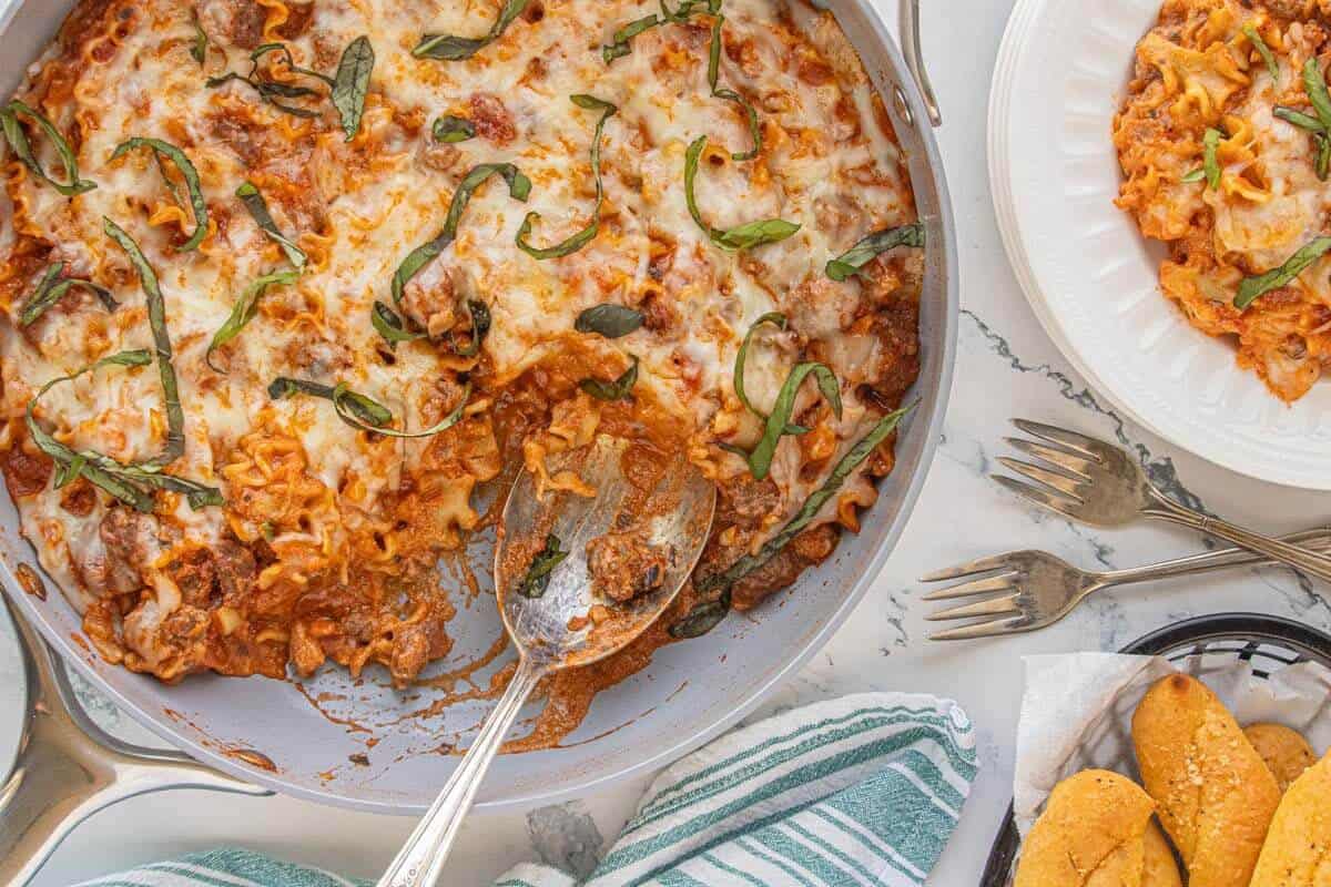 Top-down view of a skillet filled with lasagna hamburger helper, sprinkled with basil and served alongside breadsticks and a plate of pasta.