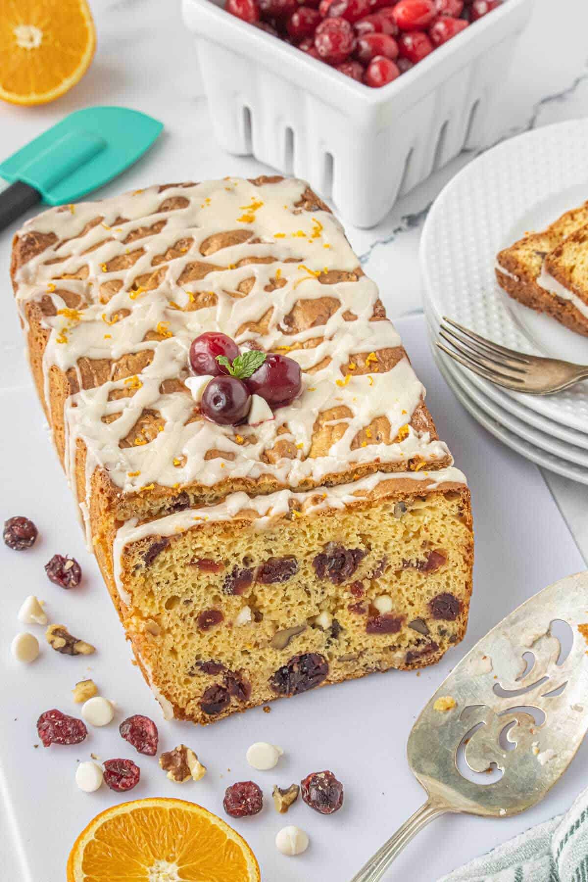 A glazed white chocolate loaf dotted with dried cranberries and white chocolate chips, topped with fresh cranberries and orange zest on a white cutting board.