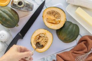 Halved acorn squash on a cutting board with seeds being scooped out using a spoon, surrounded by a knife, whole squash, butter dish, and brown striped kitchen towel.