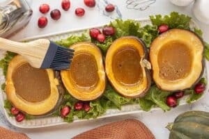 Cooked acorn squash on a platter. The center of each squash is filled with a buttery sweet sugar. The top of one squash is being brushed with the butter mixture.