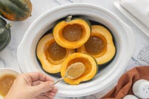 Acorn squash halves arranged cut-side up in a slow cooker, as a spoonful of brown sugar butter mixture is added to the center of one squash.