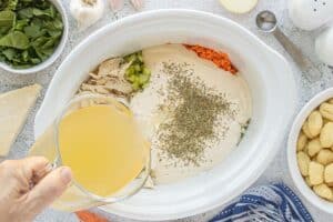 Hand pouring chicken broth into a slow cooker filled with shredded chicken, celery, shredded carrots, cream soup, and Italian herbs.