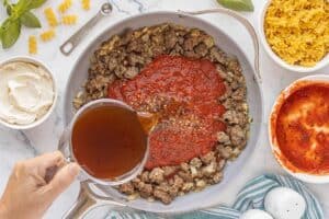 Pouring beef broth into a skillet filled with browned sausage, onions, and marinara sauce.