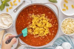 Uncooked mini lasagna noodles being stirred into a skillet of tomato meat sauce with cheese and basil nearby.