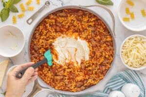Adding a scoop of ricotta cheese into a skillet of cooked noodles, beef, and tomato sauce.