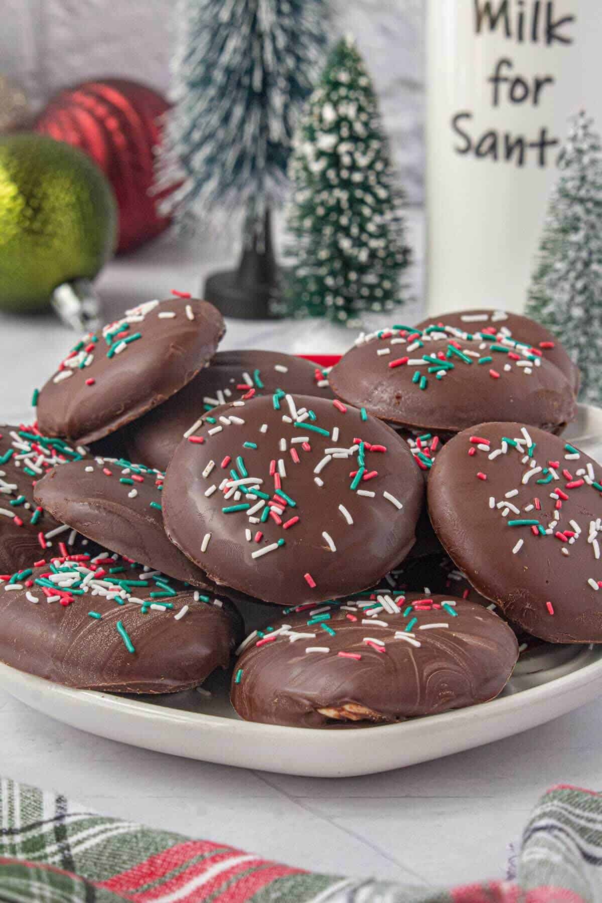 A plate of chocolate-dipped peppermint patties with colorful holiday sprinkles, surrounded by small Christmas decorations and a plaid towel for a festive backdrop.