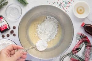 A spoonful of powdered sugar being added to a mixing bowl filled with sweetened condensed milk. Around the bowl are ingredients like peppermint extract, chocolate chips, and festive sprinkles, creating a cozy holiday baking scene.