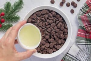 A bowl full of chocolate chips being prepared for melting, as a hand pours in a small cup of vegetable oil to create a smooth chocolate coating.