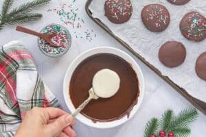 A hand dipping a round peppermint filling into melted chocolate, with a tray of already-coated candies and a jar of holiday sprinkles nearby.