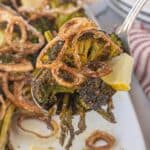 Spoon lifting a serving of roasted broccoli and asparagus topped with crispy shallots and a lemon wedge.