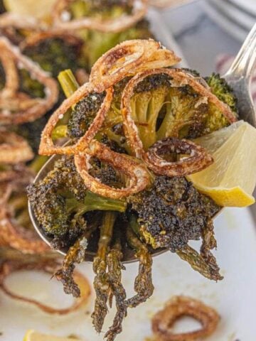Spoon lifting a serving of roasted broccoli and asparagus topped with crispy shallots and a lemon wedge.
