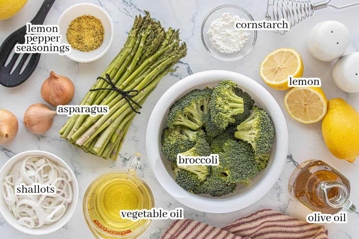 Overhead view of ingredients for roasted broccoli and asparagus, including fresh asparagus, broccoli florets, shallots, lemon, oil, corn starch, and seasonings on a marble surface.