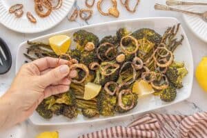 Hand scattering crispy shallots over a platter of roasted broccoli and asparagus garnished with lemon wedges and sprinkled with salt.