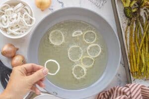 Hand placing a coated shallot ring into a pan of hot oil while other shallots fry to golden crispness.