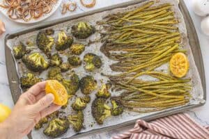 Sheet pan lined with parchment paper showing roasted broccoli florets and asparagus spears with hand squeezing roasted lemons over the vegetables.