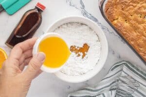 Orange juice being poured into a bowl of powdered sugar to make glaze, with a baked loaf, vanilla extract, and fresh oranges in the background.