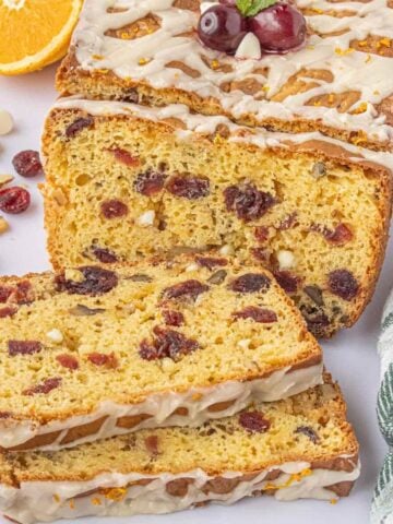 Slices of white chocolate cranberry bread showing the tender crumb with dried cranberries, white chocolate chips, and walnuts, arranged beside the glazed loaf.