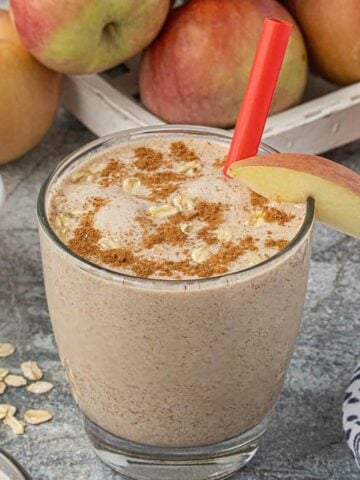 A rich apple smoothie in a glass, served with a straw. Surrounded by ingredients, like apples, oats and honey.