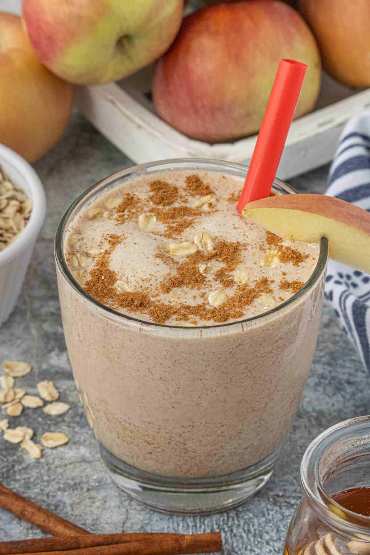 A glass filled with apple cinnamon oat smoothie, served with a straw and topped by a dash of cinnamon and sprinkled with rolled oats.