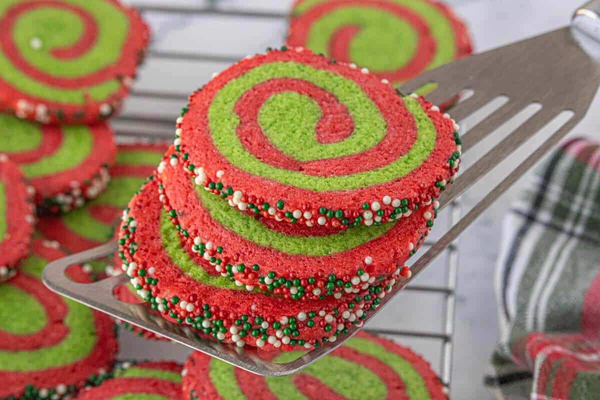 Freshly baked red and green pinwheel cookies on a spatula above a cooling rack, with sprinkle-coated edges.