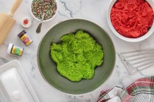Bright green cookie dough in a large bowl with a separate bowl of red dough nearby, plus food coloring jars and Christmas sprinkles on a marble counter.