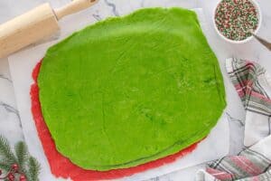 A sheet of green cookie dough placed on top of a sheet of red dough, both rolled out evenly on parchment paper with sprinkles in the background.