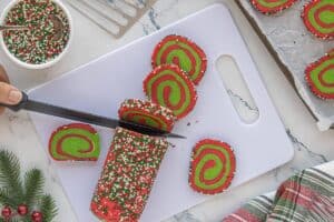 A knife slicing the chilled sprinkle-coated dough log into pinwheel cookies, revealing the red and green swirl pattern.