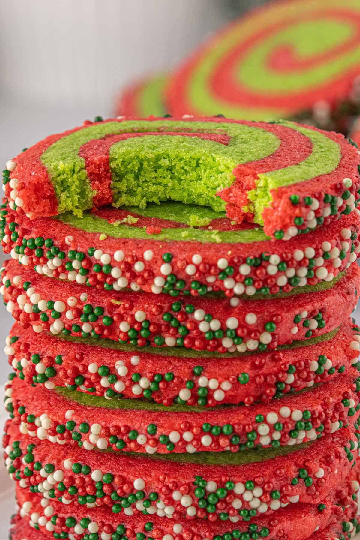 A close-up of a stack of sprinkle-coated pinwheel cookies with one cookie showing a bite taken out to reveal the soft green center.