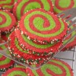 Bright red and green Christmas pinwheel cookies stacked on a metal spatula, coated with festive nonpareil sprinkles around the edges.